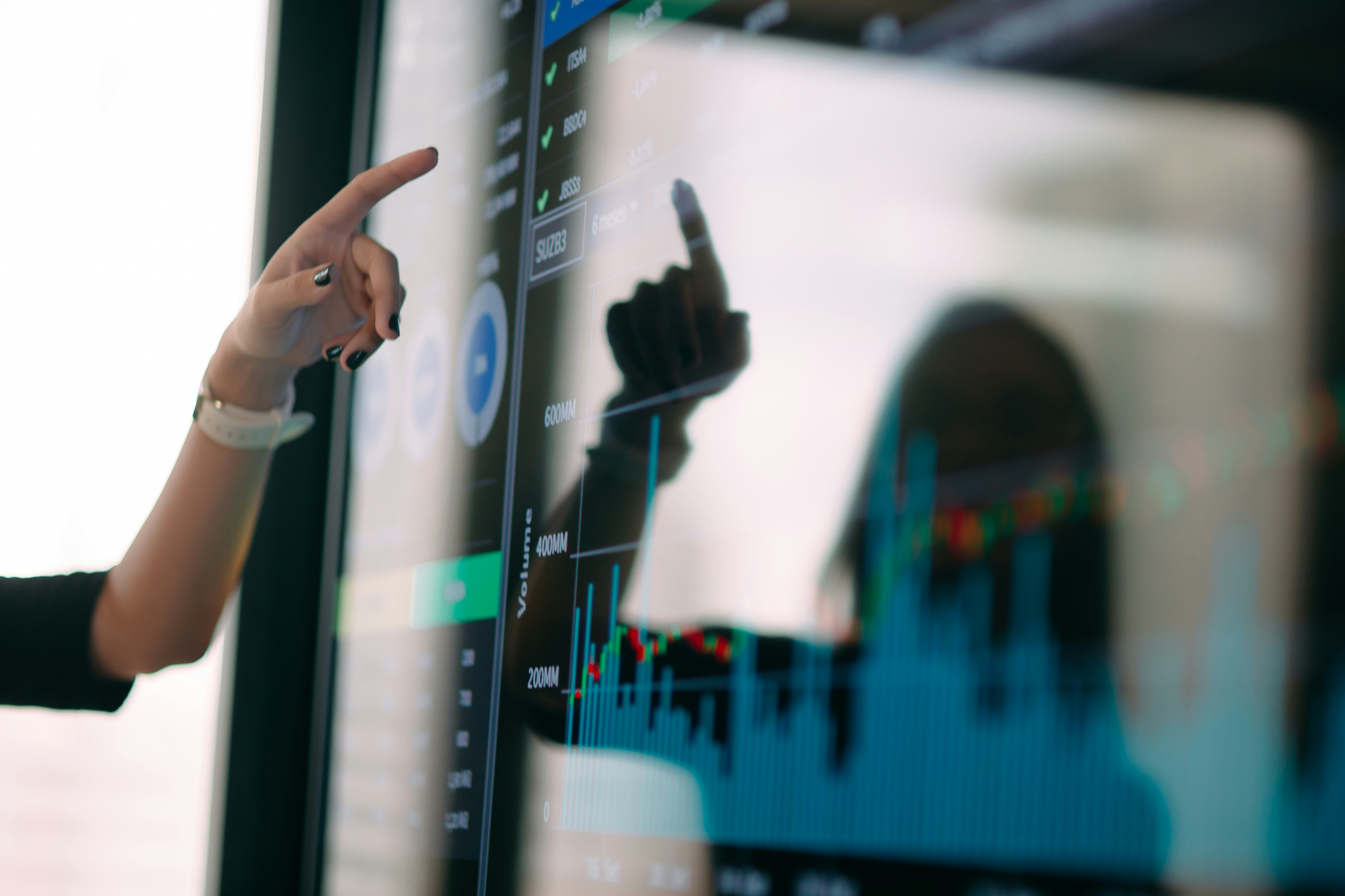 Un primo piano della mano di una persona che punta verso un grande schermo digitale che mostra grafici e dati finanziari, con il suo riflesso visibile sulla superficie lucida.